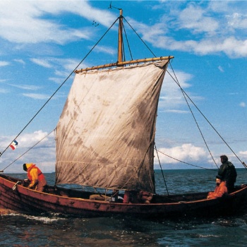 Ein kleines, aus Holz gebautes Boot gleitet über das Wasser. Das Segel ist am Mast aufgespannt und wölbt sich nach vorne. Im Heck sitzt ein Mann, ein anderer steht daneben und steuert das Schiff. Im Bug hält ein Mann einen Anker in der Hand. (© LAKD M-V/LA)