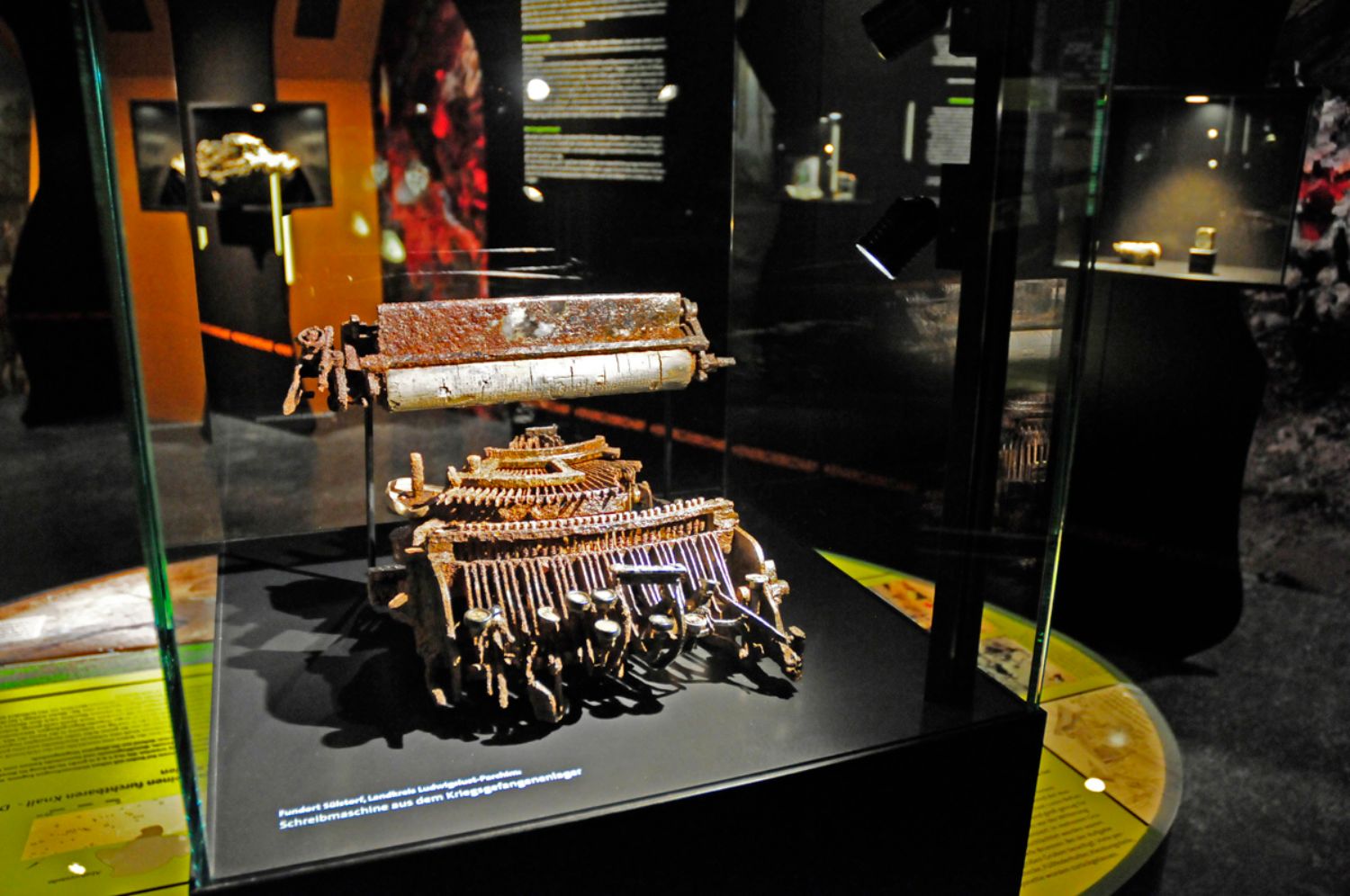 Foto aus der Sonderausstellung "PIPELINE:ARCHÄOLOGIE": In einer Vitrine steht eine verrostete Schreibmaschine. Im Hintergrund sind Ausstellungstafeln, Vitrinen und andere Ausstellungsstücke zu sehen. (© LAKD M-V/LA)
