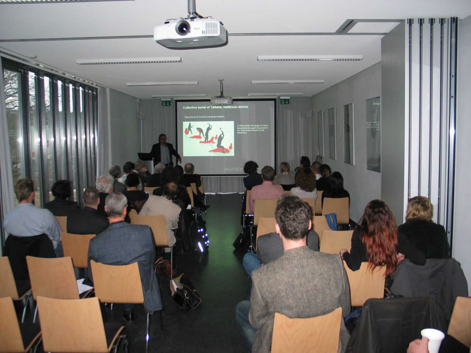 In einem Raum sitzen Menschen auf Stühlen. Vorne steht ein Redner, auf einer Leinwand ist ein Bild zu sehen. (© LAKD M-V/LA)