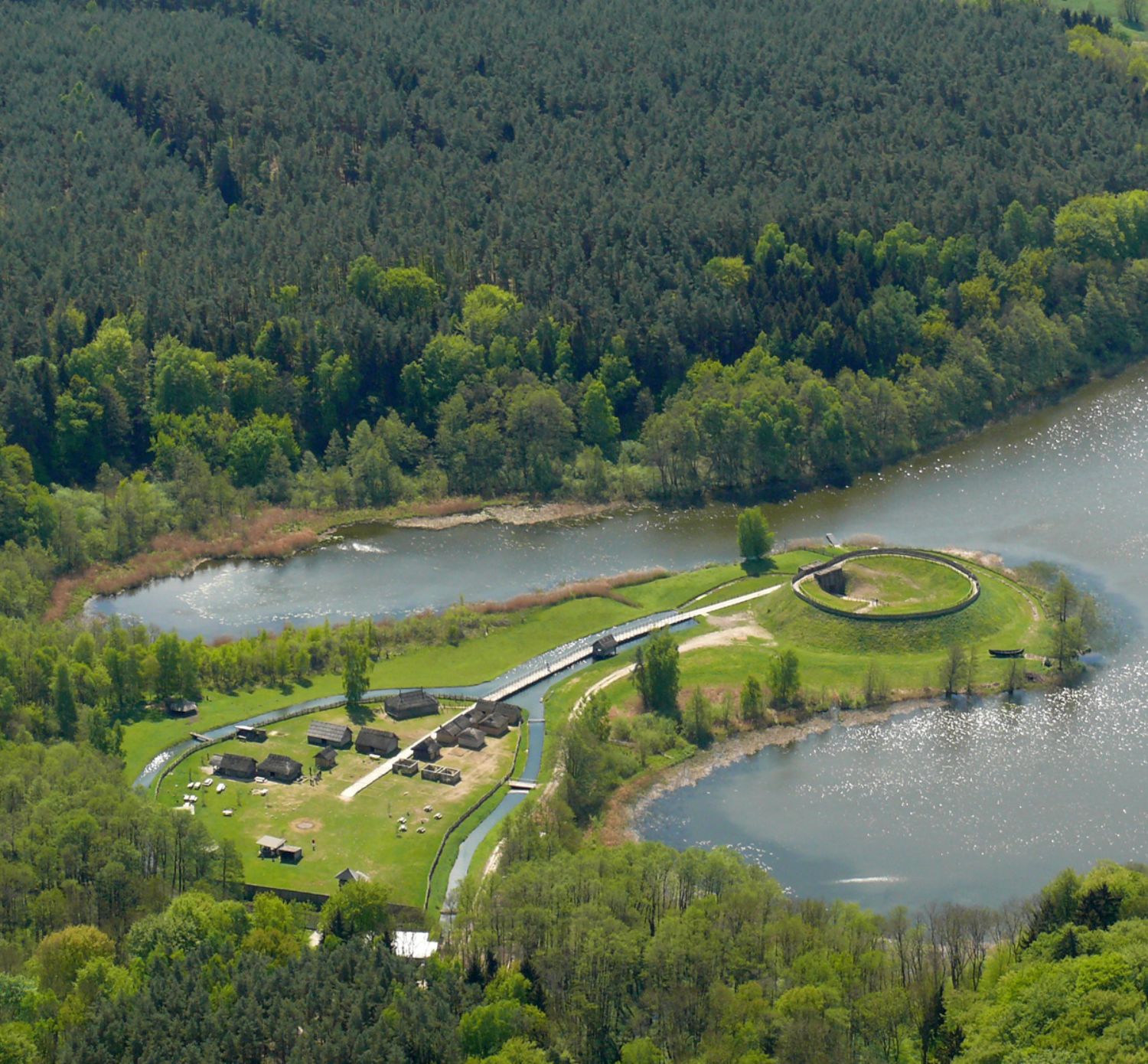 Luftbild: Eine Halbinsel, die in einen See ragt, ist aus der Vogelperspektive zu sehen. Auf der Halbinsel stehen kleine Häuser, zwischen denen ein Weg verläuft. Der Weg führt über eine Brücke zu einem ringförmigen Erdwall an der Spitze der Halbinsel. (© LAKD M-V/LA)