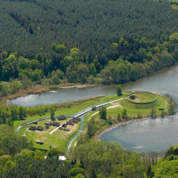 Luftbild: Eine Halbinsel, die in einen See ragt, ist aus der Vogelperspektive zu sehen. Auf der Halbinsel stehen kleine Häuser, zwischen denen ein Weg verläuft. Der Weg führt über eine Brücke zu einem ringförmigen Erdwall an der Spitze der Halbinsel. (© LAKD M-V/LA)