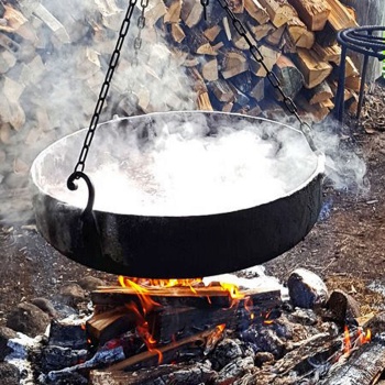 Über einem mit Steinen eingefassten Lagerfeuer hängt ein flacher, eiserner Kessel, in dem sich eine dampfende Flüssigkeit befindet. Im Hintergrund liegt Feuerholz. (© LAKD M-V/LA)