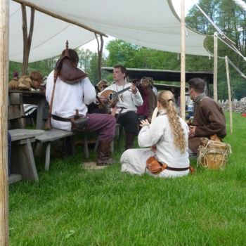 Unter einem Sonnensegel sitzen Männer und Frauen in historischen Gewändern. Ein Mann spielt auf einer Laute, ein anderer bläst auf einem Dudelsack. (© LAKD M-V/LA)