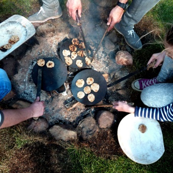 Blick von oben auf ein Lagerfeuer, an dem drei Menschen sitzen. In ihren Händen halten sie Holzstäbe, mit denen sie kleine Fladenbrote, die auf Scheiben aus dunklem Ton gebacken werden, umdrehen. Der Mann links hält einen Holztrog in der linken Hand, in dem einige fertig gebackene Fladenbrote liegen. (© LAKD M-V/LA)