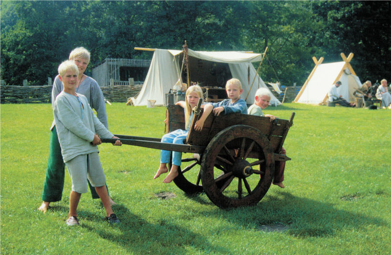 Drei Kinder sitzen auf einem zweirädrigen Wagen mit Speichenrädern. Zwei andere Kinder ziehen den Wagen. (© LAKD M-V/LA)