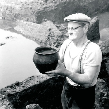 Ein Mann hält ein wassergefülltes Tongefäß in den Händen. Im Hintergrund ist eine Ausgrabungsfläche zu sehen, in der Wasser steht. (© LAKD M-V/LA)