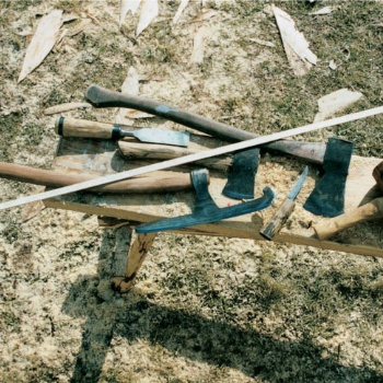 Auf einer Holzbank liegen mehrere Werkzeuge aus Eisen: Ein breiter Stechbeitel, zwei Äxte, ein Messer, eine Axt mit breit ausgezogener Schneide und ein Holzschlegel. (© LAKD M-V/LA)