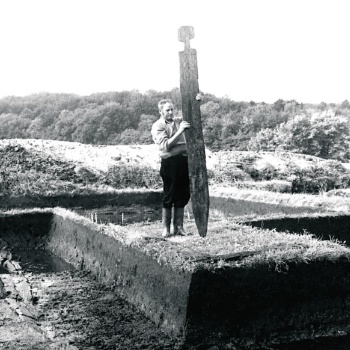 Auf einem Erdsockel zwischen Grabungsflächen steht ein Mann, der eine große, aufrecht stehende Holzbohle festhält. Die Bohle ist unten angespitzt, ihr oberes Ende ist wie ein menschlicher Kopf geformt. (© LAKD M-V/LA)