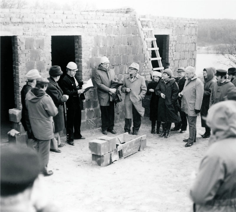 Vor dem Rohbau eines Gebäudes steht eine Gruppe von Menschen, die einen Kreis um einen Maurerkübel und einige Betonsteine bilden. Ein Mann in dunkler Arbeitskleidung und mit weißem Helm liest etwas aus einer Mappe vor. Mehrere Menschen halten etwas in der rechten Hand, wahrscheinlich ein Glas oder einen Becher. (© LAKD M-V/LA)