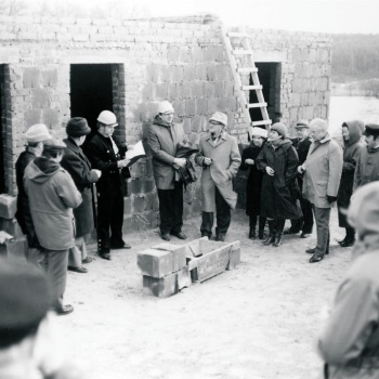 Vor dem Rohbau eines Gebäudes steht eine Gruppe von Menschen, die einen Kreis um einen Maurerkübel und einige Betonsteine bilden. Ein Mann in dunkler Arbeitskleidung und mit weißem Helm liest etwas aus einer Mappe vor. Mehrere Menschen halten etwas in der rechten Hand, wahrscheinlich ein Glas oder einen Becher. (© LAKD M-V/LA)