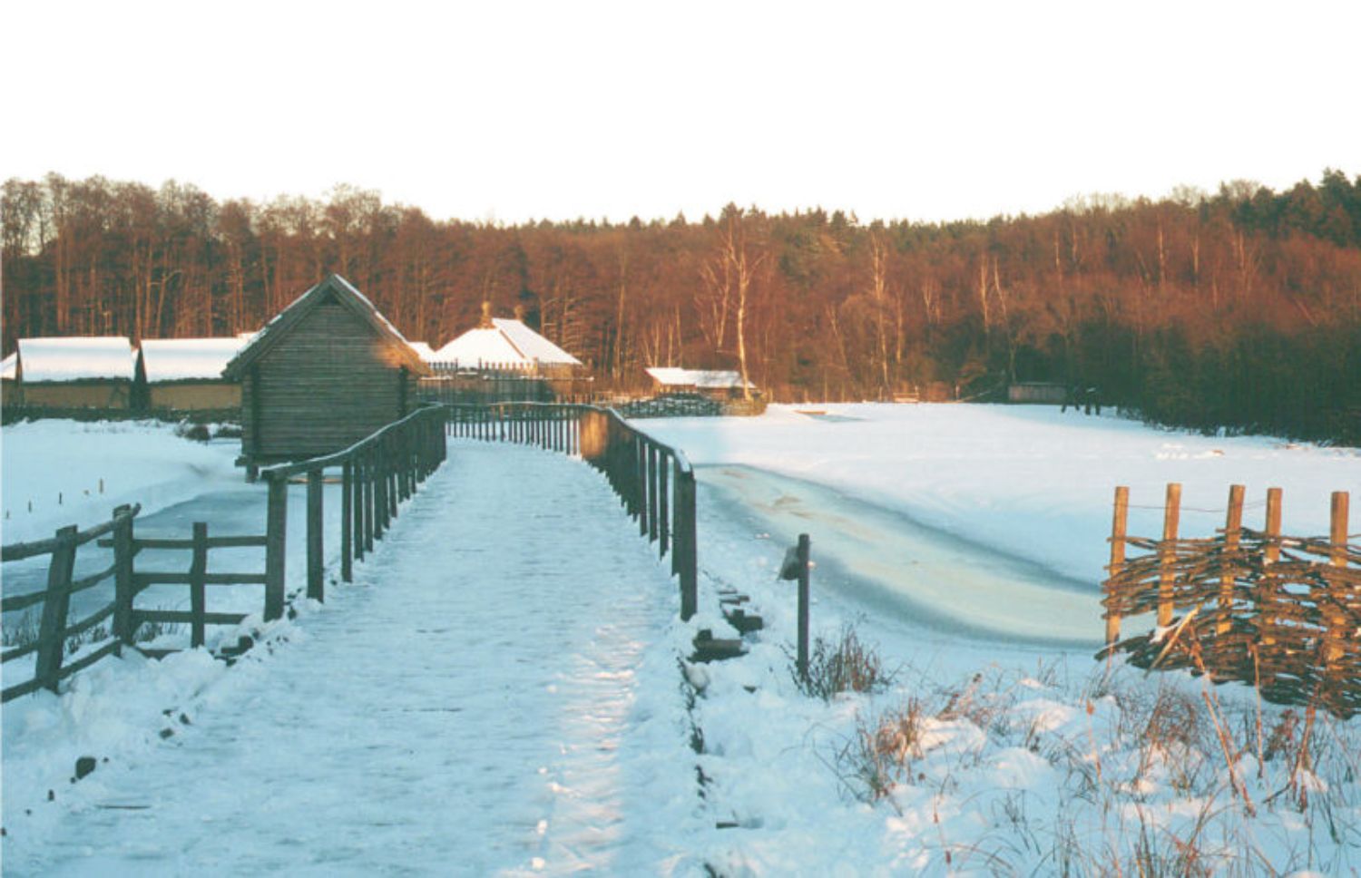 Die Landschaft ist von Schnee bedeckt. Hinter einer hölzernen Brücke, an der sich ein kleines Haus mit Holzwänden und Strohdach befindet, sind weitere Häuser mit schneebedeckten Dächern zu sehen. (© LAKD M-V/LA)