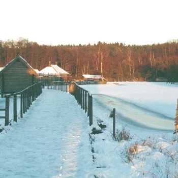 Die Landschaft ist von Schnee bedeckt. Hinter einer hölzernen Brücke, an der sich ein kleines Haus mit Holzwänden und Strohdach befindet, sind weitere Häuser mit schneebedeckten Dächern zu sehen. (© LAKD M-V/LA)