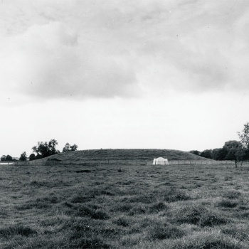 Über eine Wiese fällt der Blick auf einen grasbewachsenen Erdhügel. Links und rechts davon ist Wasser zu erahnen. Vor dem Erdhügel steht, umgeben von einem Zaun, ein weißes Zelt. (© LAKD M-V/LA)