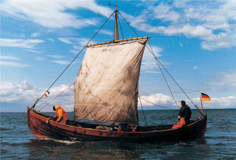 Ein kleines, aus Holz gebautes Boot gleitet über das Wasser. Das Segel ist am Mast aufgespannt und wölbt sich nach vorne. Im Heck sitzt ein Mann, ein anderer steht daneben und steuert das Schiff. Im Bug hält ein Mann einen Anker in der Hand. (© LAKD M-V/LA)