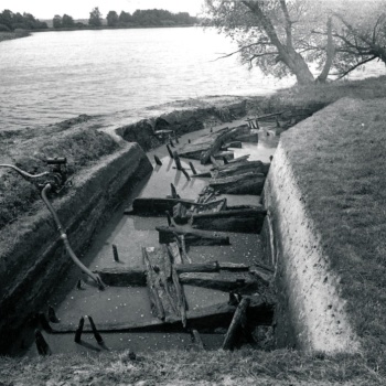 In einer mit Wasser gefüllten Grabungsfläche sind Holzkonstruktionen freigelegt. Am linken Rand steht eine Wasserpumpe, an die Schläuche angeschlossen sind. Im Hintergrund ist der See zu erkennen. Am Ufer stehen Bäume. (© LAKD M-V/LA)