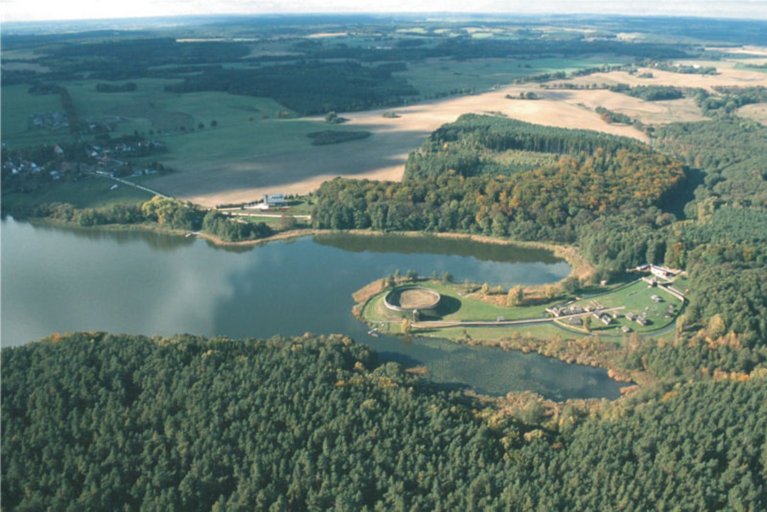 Aus der Vogelperspektive ist eine Landschaft mit Feldern, Wäldern und einem See zu sehen. Auf einer Halbinsel im See liegt ein Burgwall, im Hintergrund links das Dorf Groß Raden. (© LAKD M-V/LA)