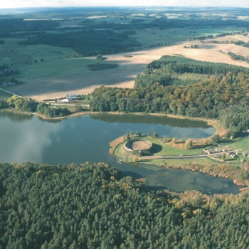 Aus der Vogelperspektive ist eine Landschaft mit Feldern, Wäldern und einem See zu sehen. Auf einer Halbinsel im See liegt ein Burgwall, im Hintergrund links das Dorf Groß Raden. (© LAKD M-V/LA)