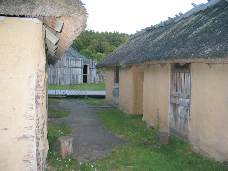 Rechts und links stehen kleine Häuser mit Lehmwänden und Strohdach, im Hintergrund ist ein großes Gebäude mit Holzwänden zu sehen. Die kleinen Häuser haben hölzerne Türen, die in der Mitte geteilt sind. Die obere Hälfte einer Tür steht offen. (© LAKD M-V/LA)