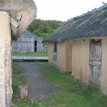 Rechts und links stehen kleine Häuser mit Lehmwänden und Strohdach, im Hintergrund ist ein großes Gebäude mit Holzwänden zu sehen. Die kleinen Häuser haben hölzerne Türen, die in der Mitte geteilt sind. Die obere Hälfte einer Tür steht offen. (© LAKD M-V/LA)