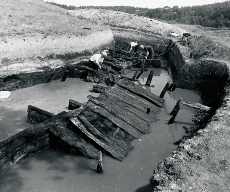 Drei Männer arbeiten in einer Grabungsfläche, in der Wasser steht. Aus dem Wasser ragen die Reste eines zur Seite gekippten Holzweges, zwischen den Männern im Hintergrund sind weitere Holzkonstruktionen zu erkennen. (© LAKD M-V/LA)