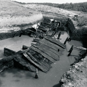 Drei Männer arbeiten in einer Grabungsfläche, in der Wasser steht. Aus dem Wasser ragen die Reste eines zur Seite gekippten Holzweges, zwischen den Männern im Hintergrund sind weitere Holzkonstruktionen zu erkennen. (© LAKD M-V/LA)