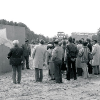 Vor einer Tafel, an der große Pläne befestigt sind, steht eine Gruppe von Menschen im Freien. Im Hintergrund liegt Baumaterial und ein hölzerner Turm ist zu sehen. (© LAKD M-V/LA)