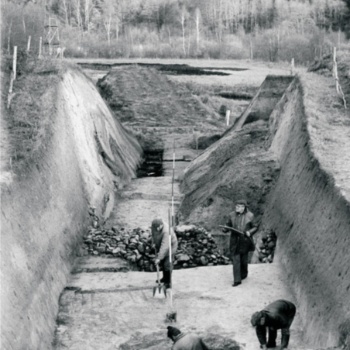 Rechts und links sind steile Erdwände zu sehen. Auf dem flachen Boden dazwischen arbeiten mehrere Männer. Einer hält ein Zeichenbrett in den Händen. Im Vordergrund kniet ein Mann in einer Grube und bearbeitet deren Rand mit einem Werkzeug. (© LAKD M-V/LA)