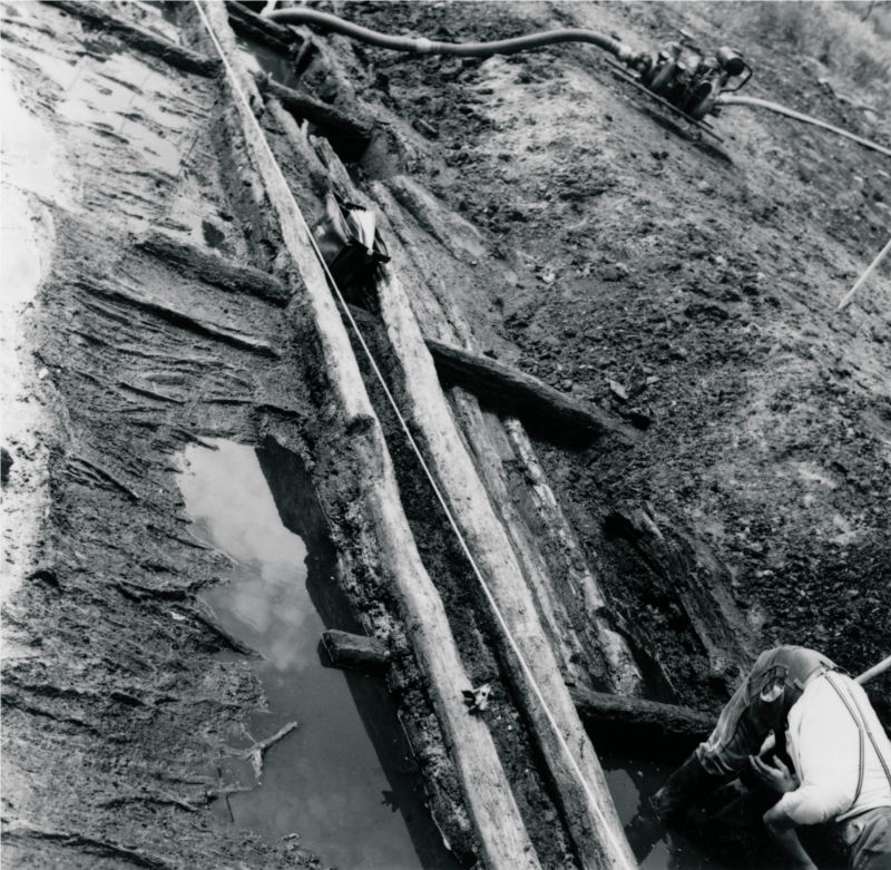 Quer durch das Bild verlaufen freigelegte Holzstämme. Daneben steht Wasser. Rechts unten ist ein Mann zu erkennen, der sich über die Hölzer beugt. Im Hintergrund steht eine Wasserpumpe mit angeschlossenen Schläuchen. (© LAKD M-V/LA)