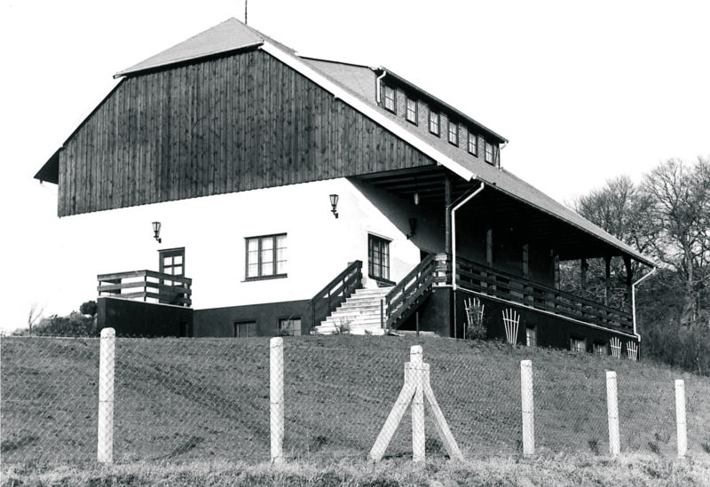 Hinter einem Maschendrahtzaun mit Betonpfählen steht ein Haus mit weißen Wänden und Walmdach. Eine Treppe führt auf eine Veranda an der Vorderseite des Hauses. (© LAKD M-V/LA)