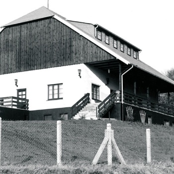 Hinter einem Maschendrahtzaun mit Betonpfählen steht ein Haus mit weißen Wänden und Walmdach. Eine Treppe führt auf eine Veranda an der Vorderseite des Hauses. (© LAKD M-V/LA)