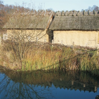 Hinter einem Wasserlauf stehen zwei kleine Häuser mit Lehmwänden und Strohdächern. Sie haben keine Fensteröffnungen. Rechts an den Häusern führt ein mit Holz belegter Weg vorbei. (© LAKD M-V/LA)