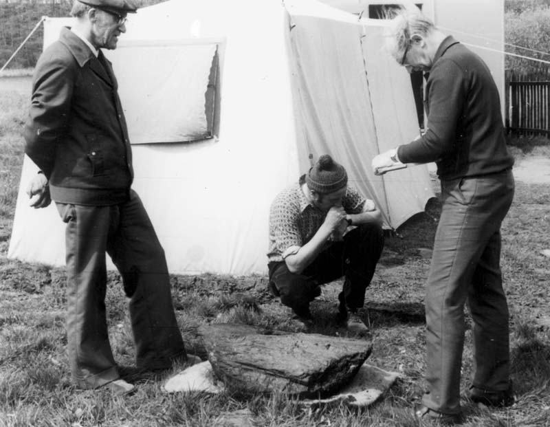 Vor einem Zelt stehen zwei Männer und sprechen miteinander. Der rechte Mann schreibt etwas auf. Zwischen ihnen kniet nachdenklich ein dritter Mann. Auf dem Boden liegt ein großes Stück Holz. © LAKD M-V/LA