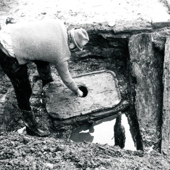 Ein Mann beugt sich über ein großes, rechteckiges Holzstück und steckt seine rechte Hand in ein Loch in der Mitte. Das Holzstück hat abgerundete Ecken und einen leicht erhöhten Rand. Rechts liegen weitere große Hölzer, links hinter dem Mann steht ein Eimer. (© LAKD M-V/LA)