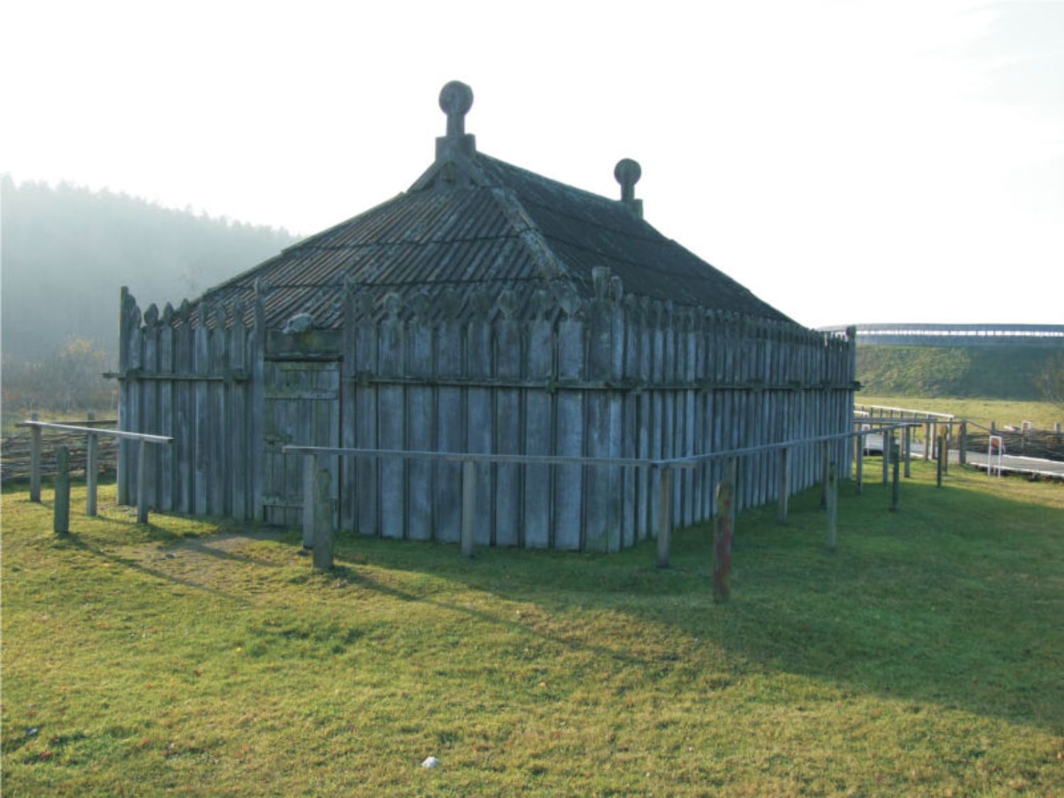 Zu sehen ist ein großes Gebäude mit hölzernen Wänden und einem aus Holz gebauten Dach. Oben aus dem Dach ragen zwei dicke, senkrecht stehende Bohlen, deren Enden wie Köpfe geformt sind. Auch die Planken der Wände haben oben Köpfe, die allerdings kleiner sind. (© LAKD M-V/LA)