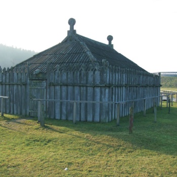 Zu sehen ist ein großes Gebäude mit hölzernen Wänden und einem aus Holz gebauten Dach. Oben aus dem Dach ragen zwei dicke, senkrecht stehende Bohlen, deren Enden wie Köpfe geformt sind. Auch die Planken der Wände haben oben Köpfe, die allerdings kleiner sind. (© LAKD M-V/LA)