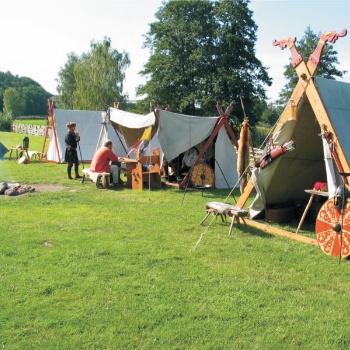 Auf einem Rasen stehen mehrere Zelte. Davor sind Schilde, ein Helm und andere Ausrüstungsgegenstände zu sehen. Ein Mensch sitzt auf einer Bank vor einem Zelt, ein anderer steht daneben, auf einen Stock gestützt. Links raucht ein kleines Lagerfeuer. (© LAKD M-V/LA )