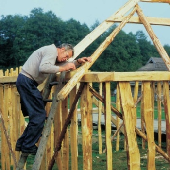 Ein Mann steht auf einer Leiter, die an einem Holzgestell lehnt. Das Holzgestell besteht aus senkrecht stehenden Pfosten, die oben durch ein waagerechtes Holz verbunden sind. Auf dem waagerechten Holz befestigt der Mann ein Rundholz, das zur Dachkonstruktion gehört. (© LAKD M-V/LA)
