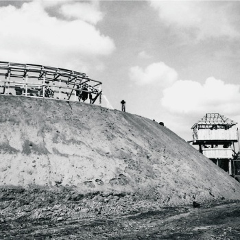 Links ist ein hoher Erdwall zu sehen, auf dem aus Holzlatten ein ringförmiges Gerüst gebaut wird. Außen an den Erdwall ist ein hölzerner Turm mit einem Walmdach angebaut. (© LAKD M-V/LA)