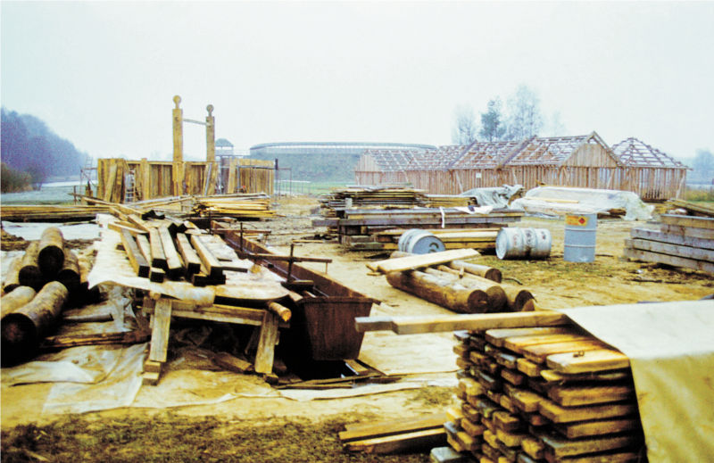 Im Vordergrund liegen Holzstapel, Fässer und anderes Baumaterial. Im Hintergrund stehen die hölzernen Gerüste mehrerer Häuser. (© LAKD M-V/LA)