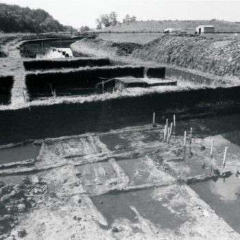 Mehrere rechteckige Grabungsflächen sind frisch ausgehoben. In ihnen steht Wasser. Dazwischen sind Holzkonstruktionen und Pflöcke zu erkennen. Im Hintergrund steht ein kleines Holzhaus vor einem grasbewachsenen Erdhügel. (© LAKD M-V/LA)