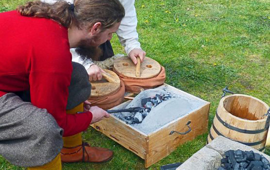 Ein Schmied, gekleidet wie ein Slawe, beugt sie über einen kleine Schmiedofen, der vor ihm auf dem Boden steht. Daneben steht eine Holzwanne mit Holzkohle. Foto: LAKD M-V/LA
