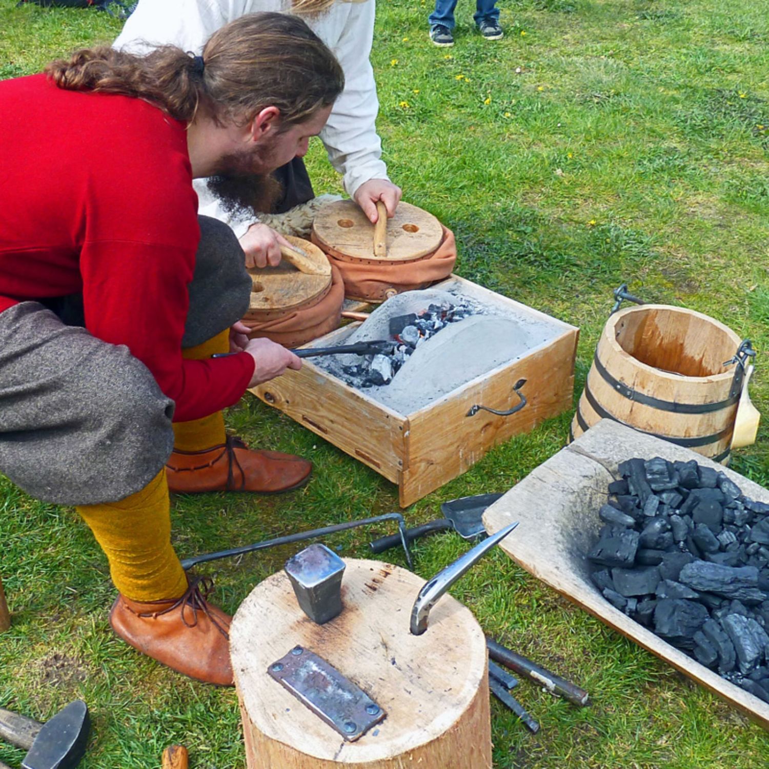 Ein Schmied, gekleidet wie ein Slawe, beugt sie über einen kleine Schmiedofen, der vor ihm auf dem Boden steht. Daneben steht eine Holzwanne mit Holzkohle. Foto: LAKD M-V/LA