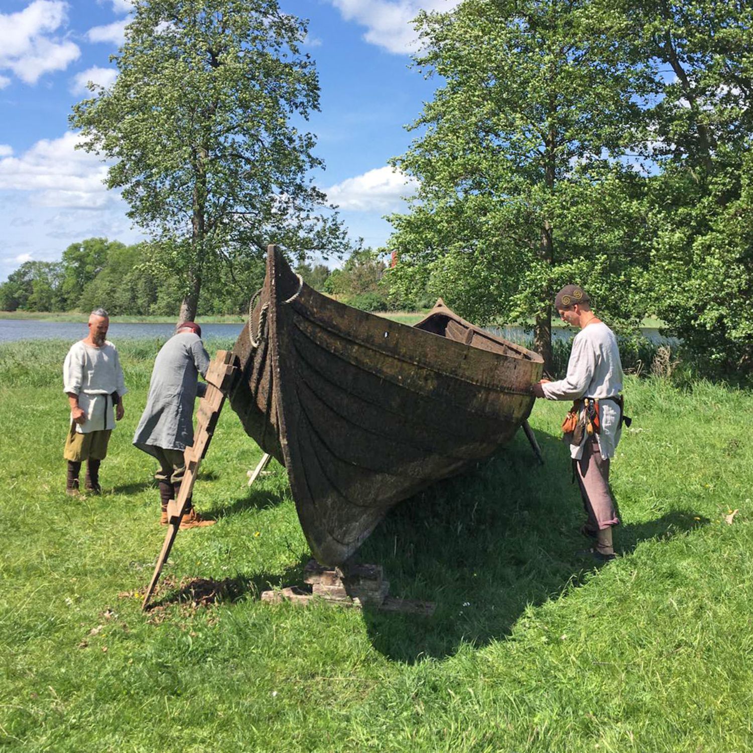 Ein Boot aus Holzplanken steht auf einer Wiese an einem See. Zwei Männer arbeiten an dem Boot, ein dritter steht daneben und betrachtet es. (© LAKD M-V/LA)