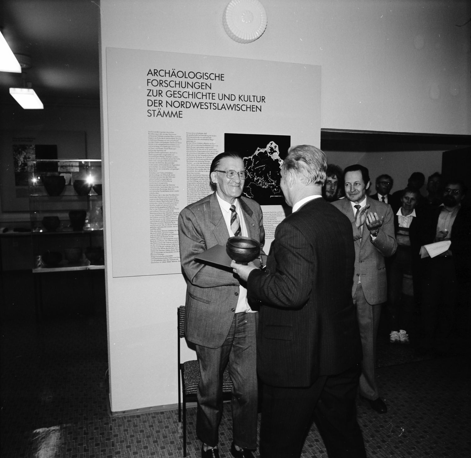 Ein Schwarz-weiß-Foto zeigt einen Mann, der vor einer Ausstellungstafel steht. Ein anderer Mann reicht ihm ein Tongefäß und schüttelt ihm die Hand. Rechts stehen Menschen und klatschen. (© LAKD M-V/LA)