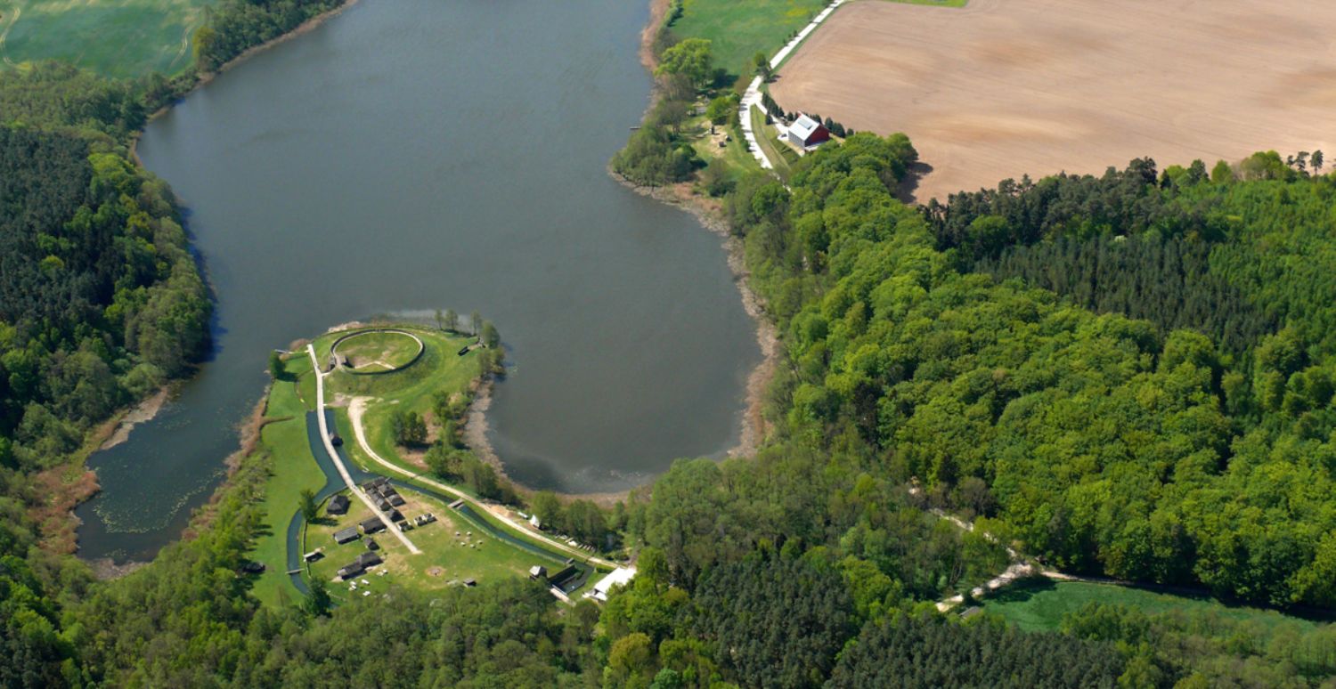 Luftbild: Aus der Vogelperspektive sind Wälder, Felder und ein See zu erkennen. In den See ragt eine Halbinsel hinein, auf der kleine Häuser stehen. An der Spitze der Halbinsel befindet sich ein ringförmiger Erdwall. An der rechten Seite des Sees steht ein Haus mit einem glänzenden Dach. (© LAKD M-V/LA)