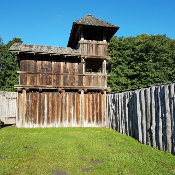 Auf einer Wiese steht ein Gebäude mit Holzwänden und einem Dach aus Holzschindeln. Der rechte Teil des Gebäudes ist dreistöckig, die beiden oberen Etagen haben große Öffnungen. Rechts an das Gebäude schließt eine Palisade aus senkrecht stehenden Eichenstämmen an. (© LAKD M-V/LA)