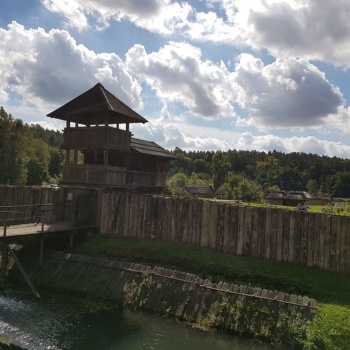 Im Vordergrund ist ein breiter Wassergraben zu sehen, dessen Böschungen mit Holzplanken befestigt sind. Über den Graben führt eine hölzerne Brücke auf ein dreistöckiges Holzgebäude zu, an das sich rechts und links eine Palisade anschließt. Hinter der Palisade stehen kleine Häuser. (© LAKD M-V/LA)