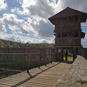 Von einer hölzernen Brücke aus fällt der Blick auf ein dreistöckiges Holzgebäude, in dessen Durchgang ein Mensch steht. Rechts und links des Gebäudes befindet sich eine Palisade aus senkrecht stehenden Holzstämmen. (© LAKD M-V/LA)