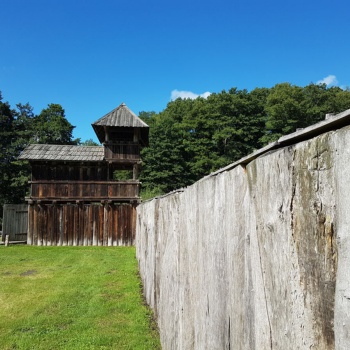 Auf einer Wiese steht ein Gebäude mit Holzwänden und einem Dach aus Holzschindeln. Der rechte Teil des Gebäudes ist dreistöckig, die beiden oberen Etagen haben große Öffnungen. Rechts an das Gebäude schließt eine Palisade aus senkrecht stehenden Eichenstämmen an. (© LAKD M-V/LA)