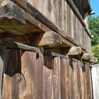 Blick auf die Seitenwand eines aus Holz gebauten Gebäudes. Das Unterteil besteht aus senkrecht stehenden Stämmen, von denen immer drei in Längsrichtung durch eine Bohle verbunden sind. Jeder vierte Stamm ragt etwas höher auf und trägt einen querliegenden Balken, der seitlich etwas über die Wand hinausragt. Darauf befindet sich ein aus Bohlen und Brettern, die mit Holznägeln verbunden sind, gebautes Stockwerk. (© LAKD M-V/LA)